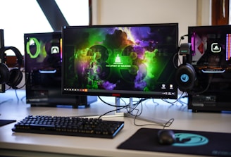 A gaming setup featuring a central monitor with a vibrant display showing 'The Spirit of Gaming'. On either side, there are gaming PCs with illuminated components and transparent cases. Headsets hang from the side of the monitor. The desk also has a black keyboard and mouse, and a branded mousepad.
