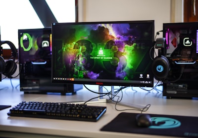 A gaming setup featuring a central monitor with a vibrant display showing 'The Spirit of Gaming'. On either side, there are gaming PCs with illuminated components and transparent cases. Headsets hang from the side of the monitor. The desk also has a black keyboard and mouse, and a branded mousepad.