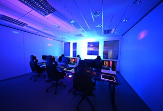A modern gaming setup with multiple high-backed gaming chairs and monitors arranged in a room with a cool blue lighting. The computers have illuminated keyboards and glowing screens, creating a high-tech ambiance.