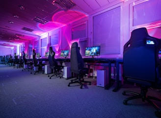 A row of gaming computers with large monitors and racing-style chairs is set up along the interior of a modern room. The space is illuminated with vibrant purple and blue LED lighting, creating a futuristic atmosphere. The room is organized and clean, suggesting a professional gaming or esports environment.