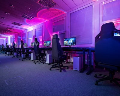A row of gaming computers with large monitors and racing-style chairs is set up along the interior of a modern room. The space is illuminated with vibrant purple and blue LED lighting, creating a futuristic atmosphere. The room is organized and clean, suggesting a professional gaming or esports environment.