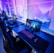 A row of modern gaming setups is arranged neatly along a window-lined wall. Each station includes a high-resolution monitor displaying vivid graphics, a mechanical keyboard with colored lighting, a gaming mouse, and an ergonomic chair. The room is illuminated with ambient blue and purple lights, creating a sleek and futuristic atmosphere.