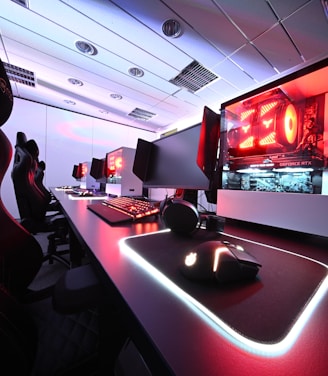 A row of high-end gaming computers on sleek desks, featuring RGB lighting and gaming accessories. The computers are housed in transparent cases with visible components, emitting a red glow. Comfortable gaming chairs with ergonomic design are positioned next to the desks, and the room's ceiling is modern with recessed lighting.