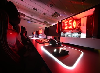 A modern gaming setup featuring several high-end computers with illuminated red interior lighting. The room is equipped with gaming chairs exhibiting a sleek black and red design. Multiple monitors are arranged on a long desk, and peripheral devices like keyboards and mice are uniformly placed, reflecting an ambiance ideal for gaming or competitive play.