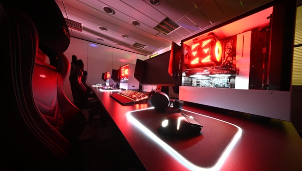 A modern gaming setup featuring several high-end computers with illuminated red interior lighting. The room is equipped with gaming chairs exhibiting a sleek black and red design. Multiple monitors are arranged on a long desk, and peripheral devices like keyboards and mice are uniformly placed, reflecting an ambiance ideal for gaming or competitive play.