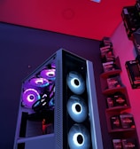A gaming computer setup featuring a high-performance PC tower with multiple RGB fans lit in blue and purple hues. The case has a modern design with transparent panels showcasing internal components. In the background, shelves are stocked with various Ryzen and other gaming components under a vibrant red and blue ambient light.