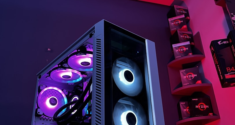 A gaming computer setup featuring a high-performance PC tower with multiple RGB fans lit in blue and purple hues. The case has a modern design with transparent panels showcasing internal components. In the background, shelves are stocked with various Ryzen and other gaming components under a vibrant red and blue ambient light.