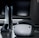 A sleek virtual reality headset sits on a reflective surface beside a gaming console and its controller. The background features a large dark monitor screen, creating a modern and futuristic setting. The lighting is subdued, highlighting the glossy surfaces of the electronic devices.