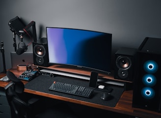 A modern gaming and streaming setup with a curved monitor placed centrally on a wooden desk. Surrounding the monitor are powerful speakers and a high-end PC tower with glowing fans. Various peripherals, including a mechanical keyboard, ergonomic mouse, and audio mixer, are neatly arranged. Overhead, a professional microphone and stand are ready for use, possibly for gaming commentary or podcasting.