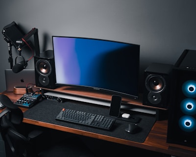 A modern gaming and streaming setup with a curved monitor placed centrally on a wooden desk. Surrounding the monitor are powerful speakers and a high-end PC tower with glowing fans. Various peripherals, including a mechanical keyboard, ergonomic mouse, and audio mixer, are neatly arranged. Overhead, a professional microphone and stand are ready for use, possibly for gaming commentary or podcasting.